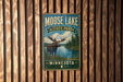 A metal sign with a moose and a lake in the background, mounted on a wooden wall.