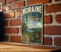 A vintage-style metal sign for Moraine Hills State Park in Illinois, featuring a scenic landscape with a trail and a sign that says "HIKE EXPLORE DISCOVER".
