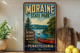 A vintage-style poster advertising Moraine State Park in Pennsylvania, featuring a scenic image of a lake with a canoe and mountains in the background.