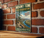 A vintage metal sign for Mount Tom State Reserve in Massachusetts, featuring a scenic mountain landscape with a river and pine trees.