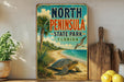 A vintage-style sign for North Peninsula State Park in Florida, featuring a beach scene with a turtle and palm trees.