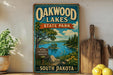 A vintage-style sign for Oakwood Lakes State Park in South Dakota, featuring a scenic landscape with a lake, trees, and mountains.