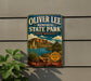 A vintage-style metal sign for Oliver Lee Memorial State Park in New Mexico, featuring a scenic landscape with mountains, a lake, and desert vegetation.