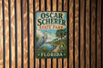 A vintage-style metal sign for Oscar Schrer State Park in Florida, featuring a blue heron and palm trees, mounted on a wooden wall.