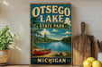 A vintage-style sign for Otsego Lake State Park in Michigan, featuring a scenic image of a lake, canoe, and forest.