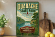a vintage-style sign for Quabache State Park, Indiana, with a scenic illustration of a river and forest. The sign is displayed on a shelf, surrounded by various kitchen items such as a cutting board, a knife, a bowl, and a potted plant.