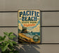 A vintage-style metal sign for Pacific Beach State Park in Washington, featuring a scenic landscape with a beach, mountains, and a pine tree.