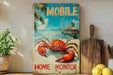 A vintage metal sign with a crab illustration and the text "MOBILE" is displayed on a shelf, accompanied by a wooden cutting board and a potted plant.
