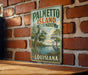 A vintage-style sign for Palmetto Island State Park in Louisiana, featuring a palm tree and a bird, is displayed on a brick wall.
