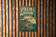 A vintage metal sign for Palm Book State Park in Michigan, featuring a tropical beach scene with palm trees and a body of water.