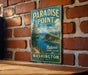 A vintage-style metal sign for Paradise Point State Park in Washington, featuring a scenic mountain landscape and a lake.