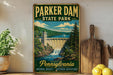 A vintage-style sign for Pennsylvania State Park, featuring a scenic illustration of a waterfall and a bridge, with the text "Parker Dam State Park" prominently displayed.