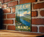 A vintage-style metal sign for Peaks- Kenny State Park in Maine, featuring a scenic landscape with mountains, pine trees, and a lake.