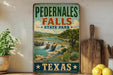 A vintage-style sign for Pedernales Falls State Park in Texas, featuring a scenic image of the waterfall surrounded by lush greenery and desert plants.