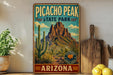 A vintage-style sign for Picacho Peak State Park in Arizona, featuring a desert landscape with mountains and cacti.