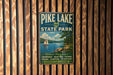 A vintage-style metal sign for Pike Lake State Park, featuring a scenic image of a lake with a sailboat, surrounded by trees and mountains.