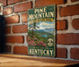 A vintage-style metal sign for Pine Mountain State Resort Park in Kentucky, featuring a scenic landscape with mountains, trees, and a lake.