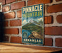 A vintage-style metal sign for Pinnacle Mountain State Park in Arkansas, featuring a scenic mountain landscape with a lake and trees.