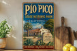 A vintage-style metal sign for Pio Pico State Historic Park in California, featuring an illustration of a Spanish-style building surrounded by palm trees and other desert vegetation.