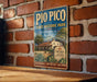 A vintage-style metal sign for Pío Pico State Historic Park in California, featuring an illustration of a Spanish-style building surrounded by palm trees and other desert vegetation.