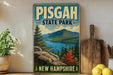 A vintage-style metal sign for Pisgah State Park in New Hampshire, featuring a scenic mountain landscape with a lake and pine trees.