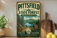 A vintage-style metal sign for the Pittsfield State Forest in Massachusetts, featuring a scenic landscape with pine trees, a lake, and mountains. The sign is displayed on a wooden shelf, with a potted plant, a wooden cutting board, and some lemons nearby.