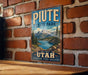 A vintage-style metal sign for Piate State Park in Utah, featuring a scenic mountain landscape and a lake.