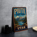 A vintage-style metal sign for Piute State Park, featuring a scenic mountain landscape and a lake, is displayed on a gray concrete surface.