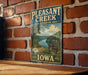 A vintage-style metal sign for the Pleasant Creek State Recreation Area in Iowa, featuring a scenic landscape with a river and forest.