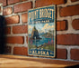 A vintage metal sign for Point Bridget State Park in Alaska, featuring a scenic landscape with mountains, a cave, and a body of water.