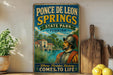 A vintage-style sign for Ponce de Leon Springs State Park in Florida, featuring a man in historical attire and a scenic illustration of the park.