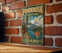 A vintage metal sign for Ponderosa State Park in Idaho, featuring a scenic mountain landscape and a lake, mounted on a brick wall.