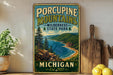 A vintage-style metal sign for Porcupine Mountains Wilderness State Park in Michigan, featuring a scenic landscape with a river, mountains, and pine trees.