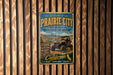 A vintage metal sign welcoming visitors to Prairie City, featuring an illustration of a vehicle and a scenic landscape.