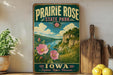A vintage-style sign for Prairie Rose State Park in Iowa, featuring a scenic landscape with a river, mountains, and pink flowers.