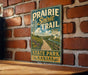 A vintage-style metal sign for the Prairie Spirit Trail in Kansas, featuring a scenic illustration of a path leading through a grassy field with a river and a blue sky.