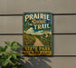 A vintage-style metal sign for the Prairie Spirit Trail in Kansas, featuring a scenic landscape with a dirt path, grassy fields, and a river.