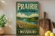 A vintage-style sign for Prairie State Park in Missouri, featuring a scenic landscape with a river, grass, and flowers.