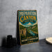 A vintage-style metal sign for Preparation Canyon State Park, featuring a scenic landscape with a river and mountains, is displayed on a gray countertop next to some black camera lenses.