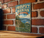 a vintage-style metal sign with a scenic mountain landscape, mounted on a brick wall.