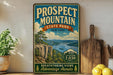 a vintage-style sign for Prospect Mountain State Park in New York, with a scenic mountain landscape and a wooden cutting board on a shelf.