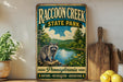 A vintage-style sign for Raccoon Creek State Park in Pennsylvania, featuring a raccoon on a rock overlooking a river surrounded by trees and mountains.