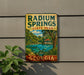 A vintage-style metal sign for Radium Springs State Park in Georgia, featuring a scenic landscape with a lake, palm trees, and a gazebo.