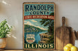 A vintage-style sign for Randolph County, Illinois, featuring a scenic landscape with a river, trees, and mountains.