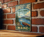 A vintage metal sign for Rangeley Lake State Park in Maine, featuring a scenic illustration of a lake, mountains, and a canoe.