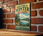 A vintage-style metal sign for Raymond B. Winter State Park in Pennsylvania, featuring a scenic landscape with a lake and mountains.
