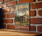 A vintage-style metal sign for Riordan Mansion State Historic Park in Arizona, featuring an illustration of a cabin surrounded by trees and mountains.
