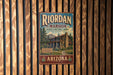 A vintage metal sign for the Riordan mansion, a historic park in Arizona, is displayed on a wooden fence.