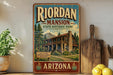 A vintage-style sign for Riordan Mansion State Historic Park in Arizona, featuring a cabin surrounded by trees and mountains.