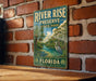 A vintage-style metal sign for River Rise Preserve, Florida, featuring a scenic illustration of a river surrounded by palm trees and a heron.
